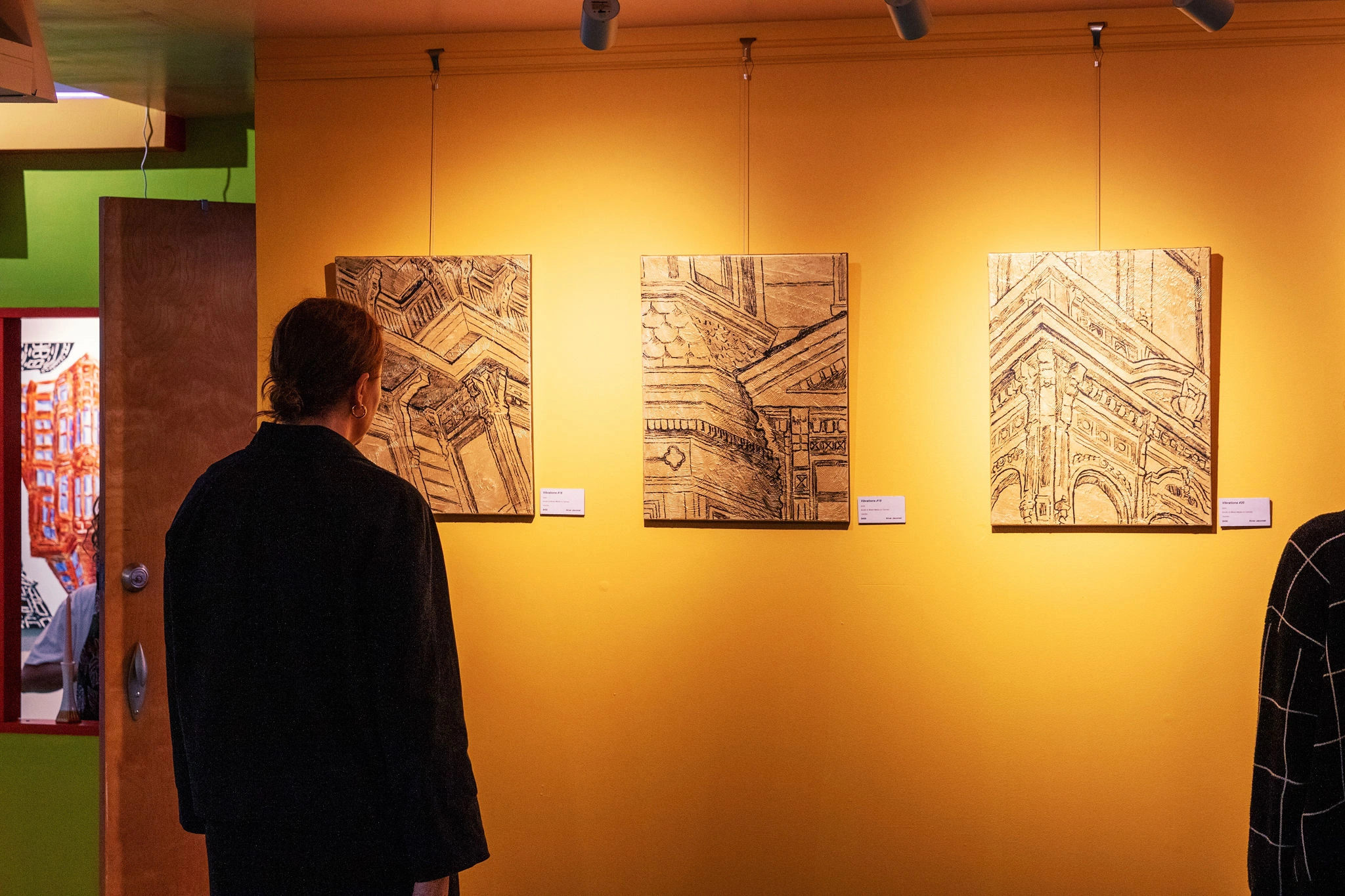 A person views three architectural drawings on a vibrant yellow wall in an art gallery, conveying a contemplative and artistic atmosphere.